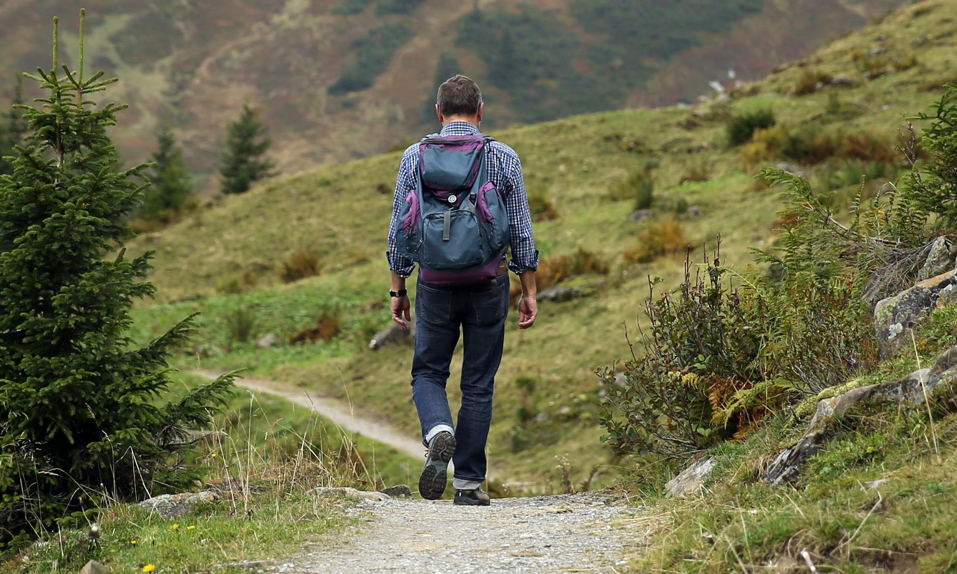 man carrying backpack walking near green tree and grass duriong daytime